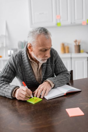 Photo for Grey haired man with azheimers syndrome writing phone number on sticky notes near blank notebook on table in kitchen - Royalty Free Image