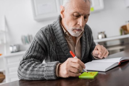 senior man with alzheimer syndrome writing on sticky note near blank notebook 