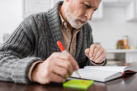 thoughtful man with alzheimer syndrome looking at blank notebook while holding blurred felt pen near sticky notes