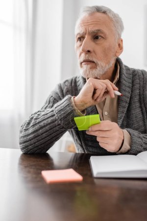 Foto de Senior man with alzhemeirs disease holding sticky notes near empty notebook and looking away in kitchen - Imagen libre de derechos