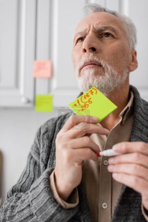 Foto de Tense man suffering from memory loss while holding sticky notes with phone number and looking away in kitchen - Imagen libre de derechos