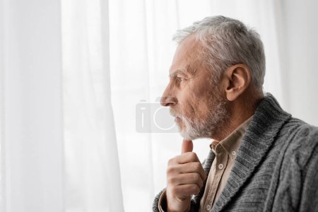 Foto de Side view of aged man suffering from memory loss and holding hand near chin while looking away at home - Imagen libre de derechos