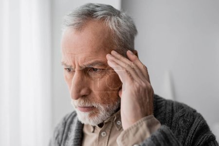 Photo for Grey haired senior man with alzheimer syndrome touching head while looking away and thinking at home - Royalty Free Image