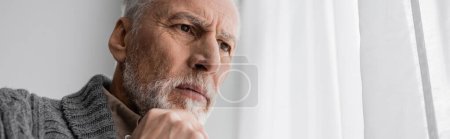 Foto de Bearded senior man with alzheimer syndrome holding hand near chin and looking away at home, banner - Imagen libre de derechos
