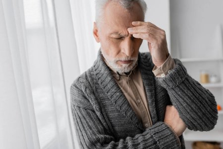 Foto de Depressed senior man with alzheimer syndrome standing with closed eyes and touching forehead at home - Imagen libre de derechos