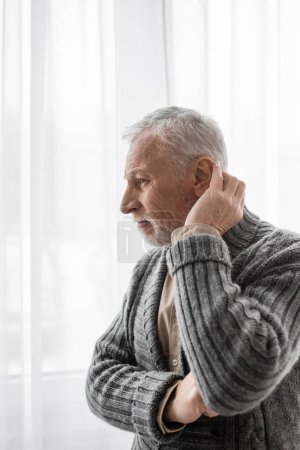 Foto de Senior and thoughtful man in knitted cardigan looking away near window while suffering from alzheimer syndrome - Imagen libre de derechos