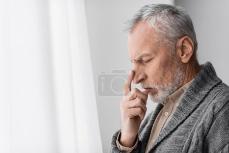 Photo for Side view of depressed senior man with alzheimer syndrome touching face while standing at home - Royalty Free Image