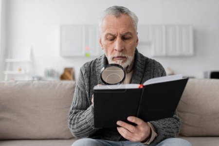 Foto de Senior man with alzheimer disease sitting on sofa and looking in notebook through magnifying glass - Imagen libre de derechos