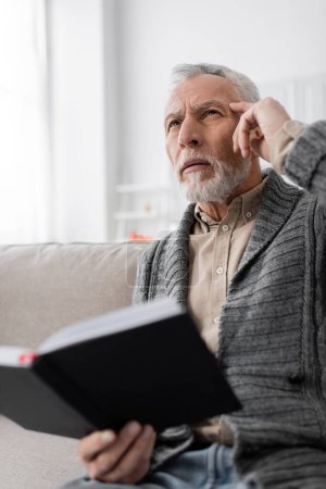 senior man suffering from memory loss and touching head while thinking on couch with blurred notebook
