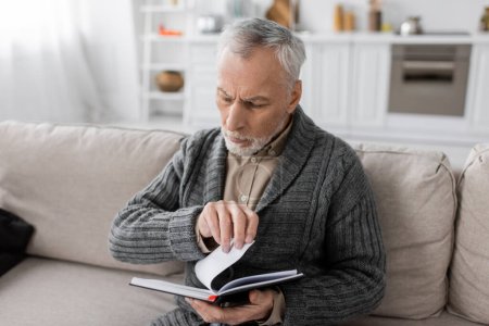grey haired man suffering from memory loss caused by alzheimer disease looking in notebook on couch at home