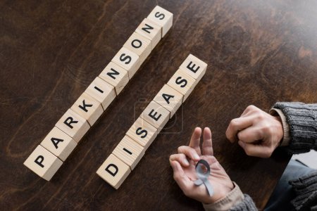 Foto de High angle view of cropped man holding grey ribbon in trembling hands cubes with parkinsons disease lettering on wooden table - Imagen libre de derechos