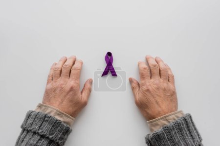 top view of hands of senior man with alzheimer syndrome near purple ribbon on white surface
