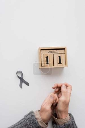 top view of grey ribbon and wooden calendar with april 11 date near cropped man with parkinson syndrome and tremor in hands on white surface
