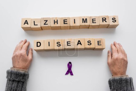 Foto de Top view of cropped senior man near purple ribbon and wooden cubes with alzheimers disease lettering on white surface - Imagen libre de derechos