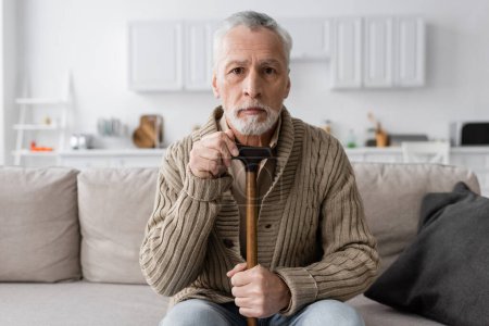 depressed man suffering from dementia while sitting with walking cane and looking at camera at home