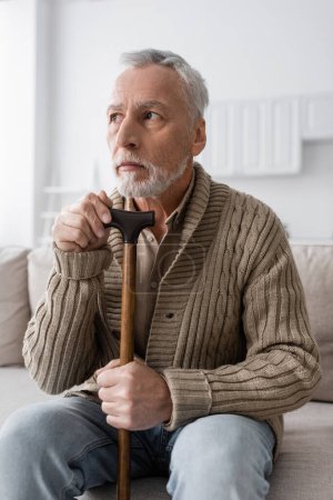 senior man with alzheimer disease looking away while sitting with walking cane at home