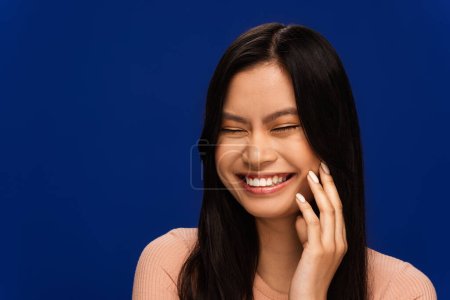 Téléchargez les photos : Cheerful asian woman touching face and closing eyes isolated on blue - en image libre de droit