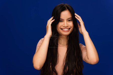 Cheerful asian woman touching long hair isolated on blue 