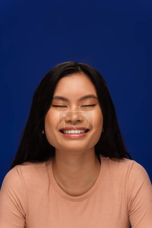 Foto de Portrait of joyful asian model closing eyes isolated on blue - Imagen libre de derechos