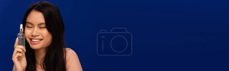Photo for Positive asian woman holding cosmetic serum isolated on blue, banner - Royalty Free Image