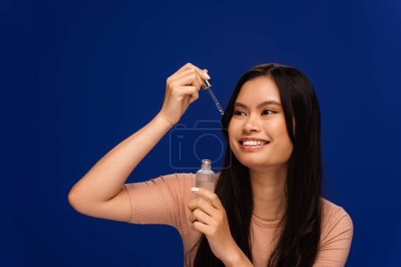 Cheerful asian woman holding cosmetic serum and looking at dropper isolated on blue 