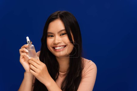 Cheerful asian woman in t-shirt holding serum isolated on blue 