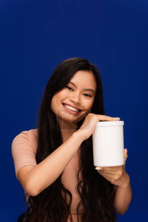 Smiling asian woman holding jar with cosmetic product isolated on blue 