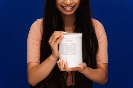 Partial view of positive woman holding jar of cosmetic product isolated on blue 
