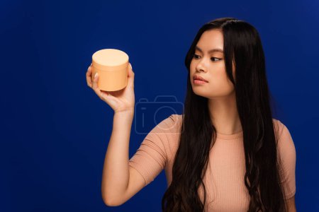 Brunette asian model looking at hair mask isolated on blue 