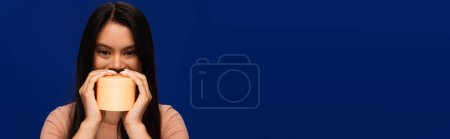 Téléchargez les photos : Young asian model holding body cream near face isolated on blue, banner - en image libre de droit