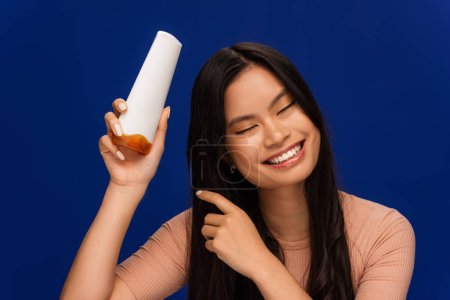 Smiling asian woman holding hair conditioner isolated on blue 