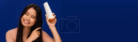 Photo for Brunette asian woman holding hair conditioner isolated on blue, banner - Royalty Free Image