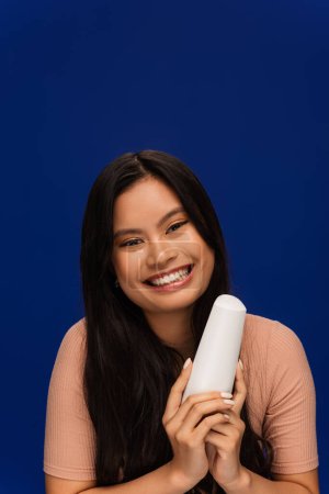 Positive asian woman in t-shirt holding hair conditioner isolated on blue 