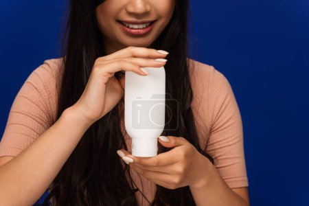 Cropped view of positive brunette woman holding cosmetic lotion isolated on blue 