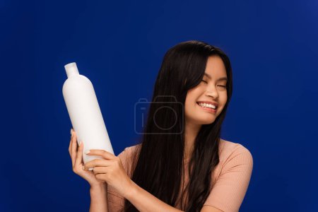 Foto de Cheerful asian woman holding bottle of shampoo isolated on blue - Imagen libre de derechos