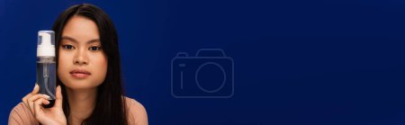 Young asian woman holding face foam and looking at camera isolated on blue, banner 