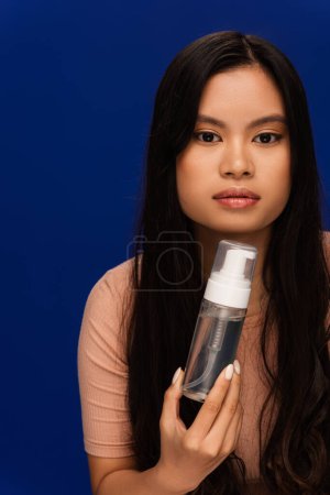 Asian woman in beige t-shirt holding face foam and looking at camera isolated on blue 