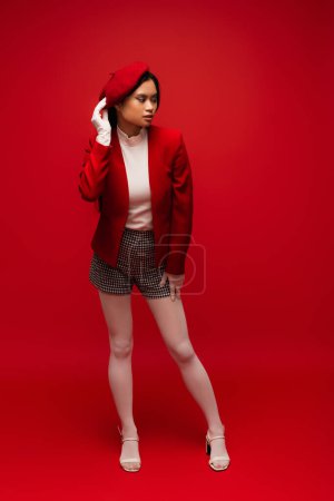 Full length of stylish asian woman in shorts and jacket standing on red background 
