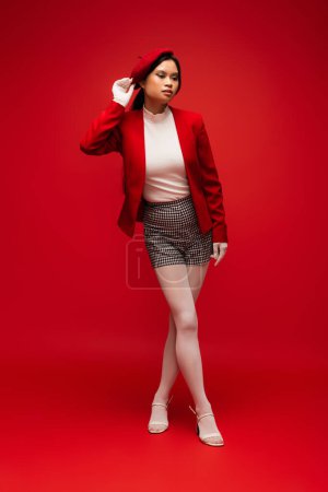 Asian woman in gloves touching beret and posing on red background 