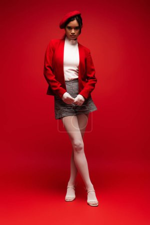 Full length of trendy asian model in tights and gloves standing on red background 