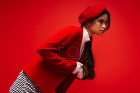 Trendy asian model in white gloves adjusting jacket isolated on red 