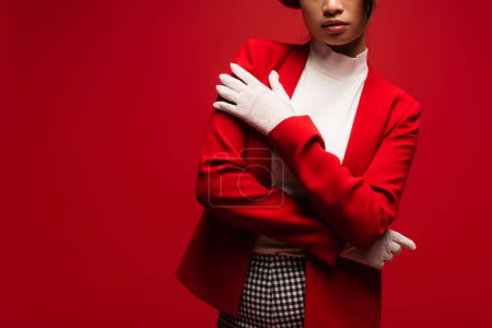 Cropped view of stylish woman in jacket and gloves posing isolated on red 