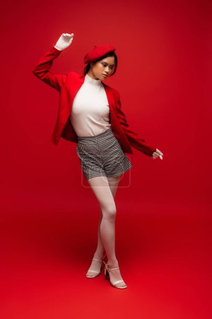 Full length of trendy asian woman in beret and blazer posing on red background 
