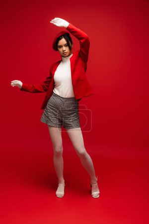 Full length of fashionable asian woman in blazer and shorts standing on red background 
