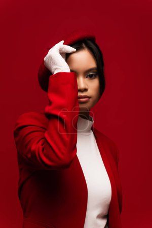 Trendy asian woman in jacket and glove adjusting beret isolated on red 