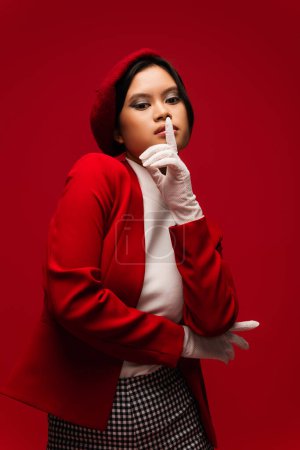 Stylish asian model in beret and jacket showing shh gesture isolated on red 