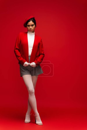 Full length of stylish asian woman in jacket and tights looking at camera on red background 