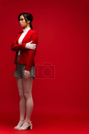 Full length of asian woman in beret and plaid posing shorts on red background 