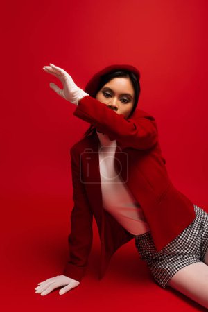 Asian woman in blazer ad white gloves looking at camera while sitting on red background 
