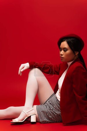 Fashionable asian model in beret and white tights sitting on red background 